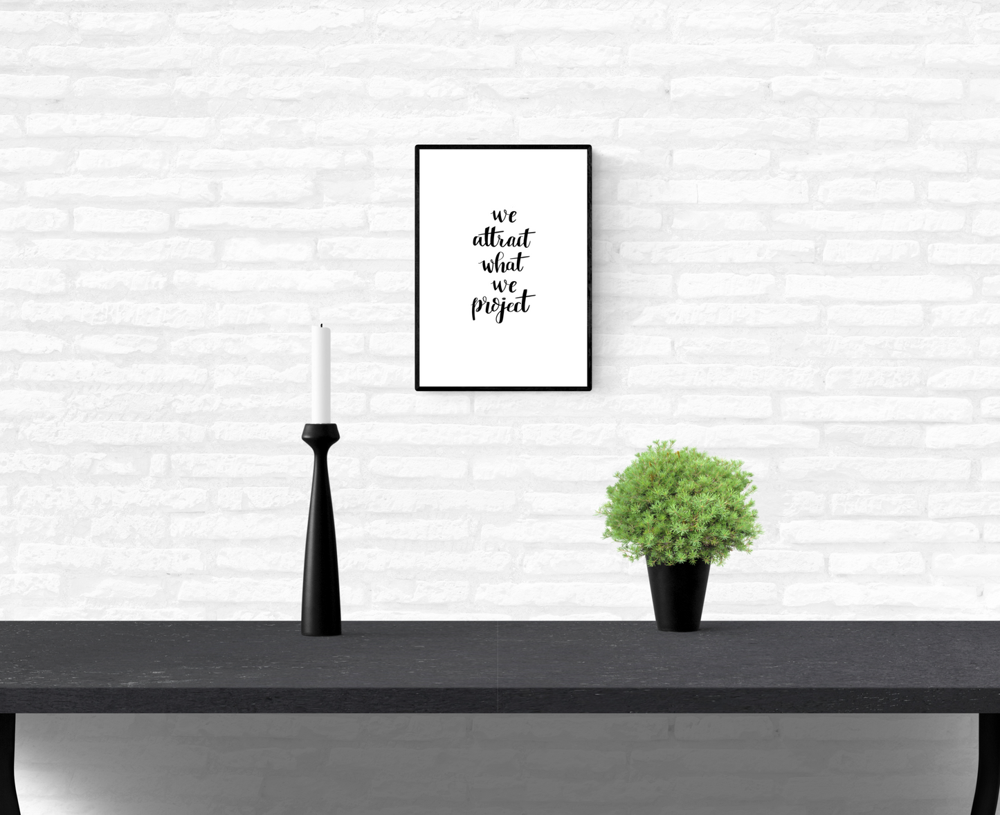 Wall quote print with the words, “we attract what we project”, framed and mounted on a home’s interior white brick wall