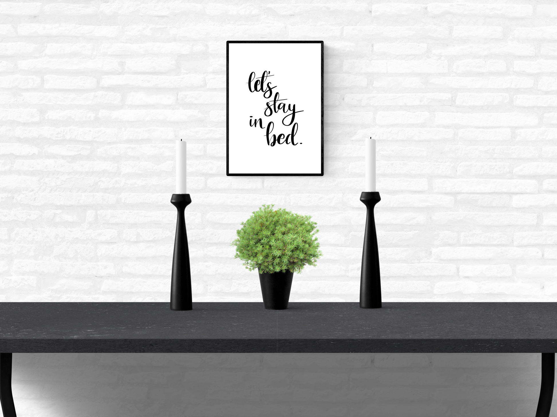 Framed wall quote typography print with the words “let’s stay in bed”, mounted on a home’s interior white brick wall above a table