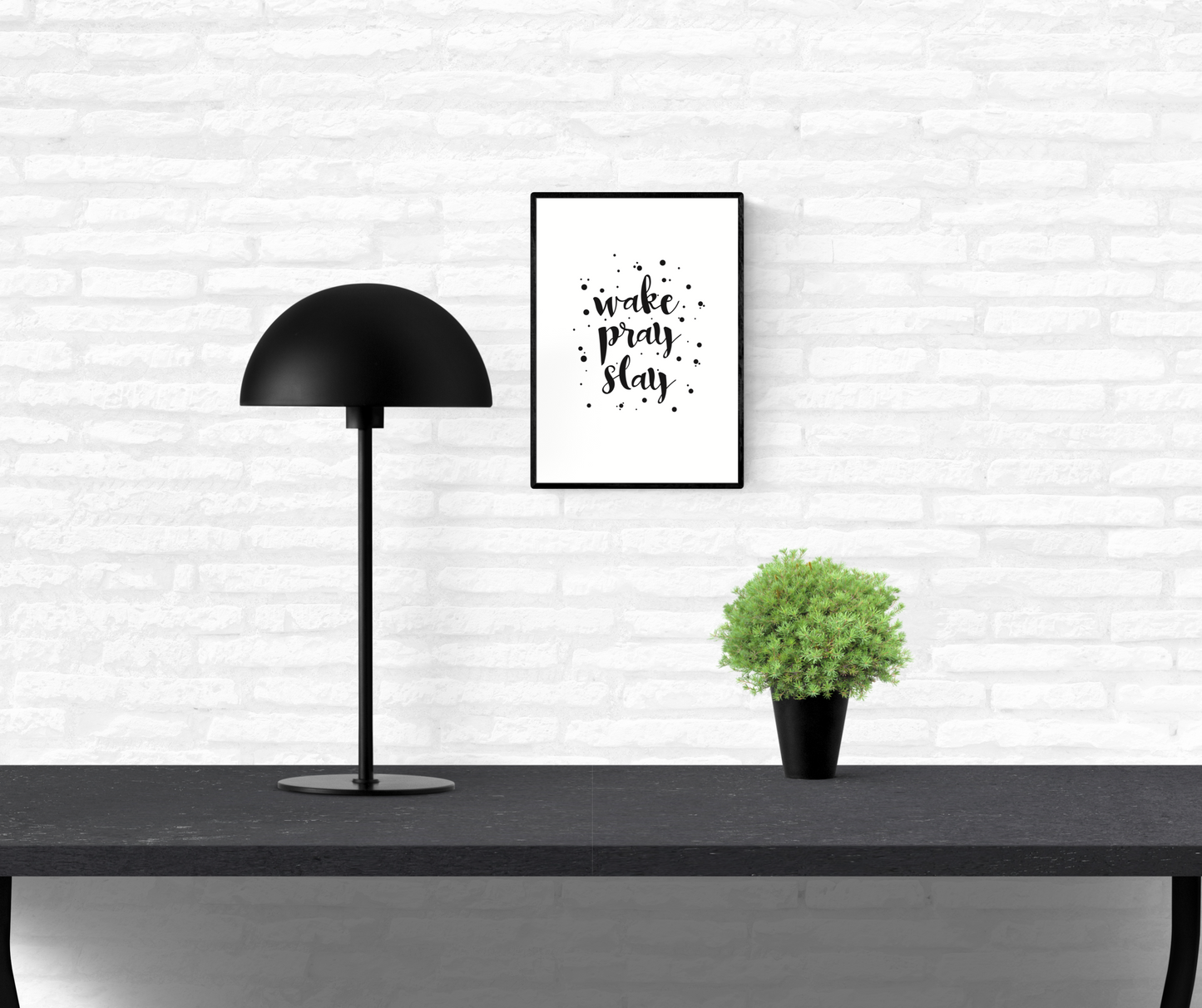 Quote wall print with the words, “wake pray slay”, surrounded by black dots, framed and mounted on an interior white brick home wall above a table
